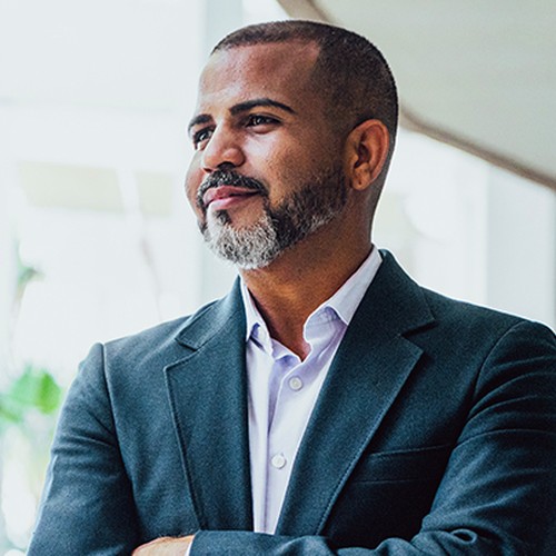 A man in a dark blazer and light shirt stands confidently with his arms crossed, gazing thoughtfully. He is positioned in a modern, bright space with plants in the background.