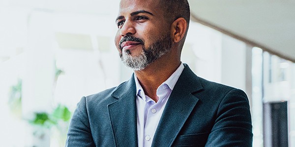 A man in a dark blazer and light shirt stands confidently with his arms crossed, gazing thoughtfully. He is positioned in a modern, bright space with plants in the background.