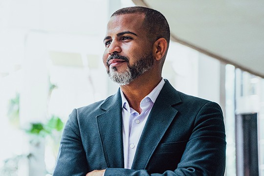 A man in a dark blazer and light shirt stands confidently with his arms crossed, gazing thoughtfully. He is positioned in a modern, bright space with plants in the background.