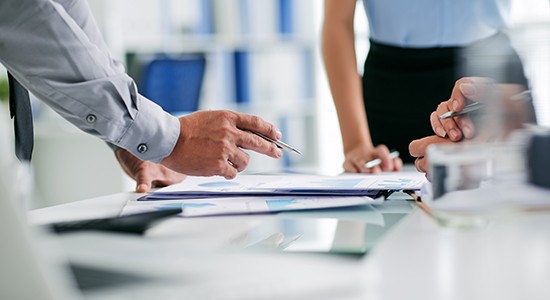 A hand with a pen gestures over documents on a table, while other hands are poised nearby, suggesting a collaborative discussion in a modern office environment.