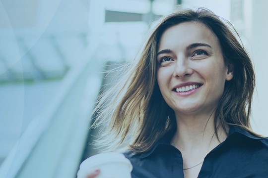 A woman with shoulder-length hair smiles while holding a coffee cup. She stands in a bright, modern indoor space with large windows, suggesting a welcoming atmosphere.