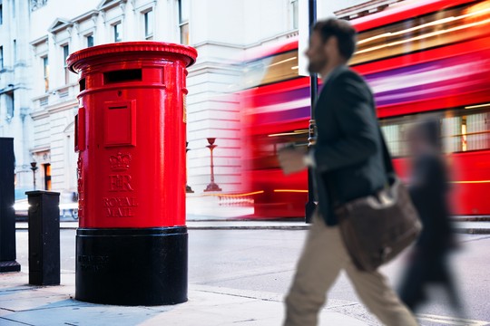 A red postbox stands on a city street, while a man walks by, holding a drink. A blurred bus speeds past in the background, suggesting a busy urban environment.