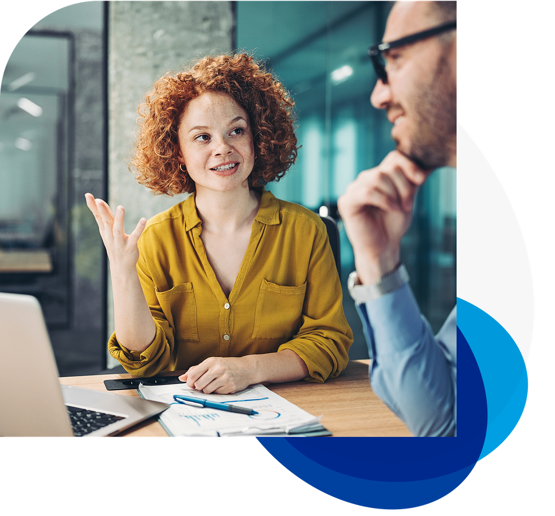 A woman with curly red hair wearing a yellow shirt gestures while speaking to a man in a blue shirt. They are sitting at a wooden table with a laptop and paperwork in a modern office setting.
