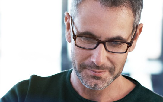 A man with glasses is focused intently on something in front of him, likely reading or working. The background shows a softly lit, modern indoor space.