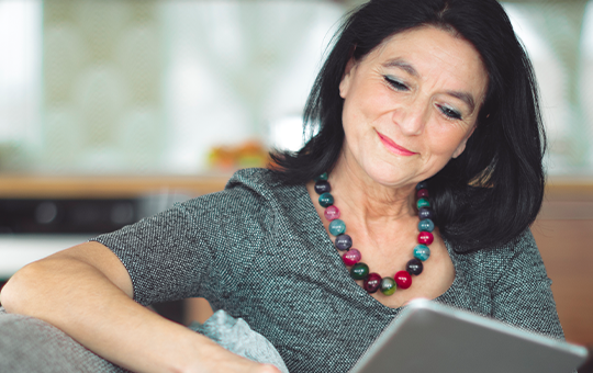A woman with long black hair, wearing a grey top and a colorful necklace, is sitting comfortably on a couch, smiling while using a tablet in a well-lit kitchen environment.