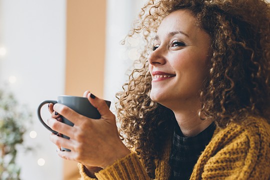 A woman with curly hair holds a mug, smiling while gazing off to the side. She is seated indoors, surrounded by soft lighting and a cozy atmosphere.