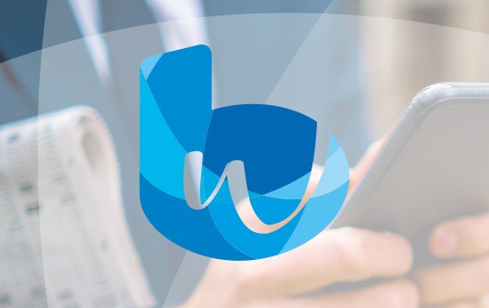 A person holds a smartphone while dressed in business attire, standing outdoors with a blurred background. A blue logo featuring stylized letters is prominently displayed in the foreground.