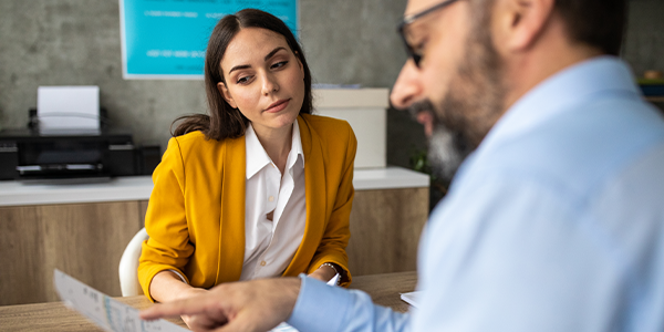 A woman in a yellow blazer listens intently as a man in glasses gestures toward papers on a table, discussing documents in a modern office environment with a printer in the background.