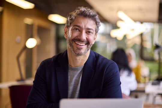 A smiling man with curly hair sits in front of a laptop, engaging in conversation within a bright, modern workspace featuring large windows and contemporary lighting fixtures.