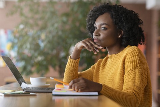 A woman in a yellow sweater sits at a table, thoughtfully resting her chin on her hand. A laptop and a white cup are nearby, surrounded by plants and colorful artwork.