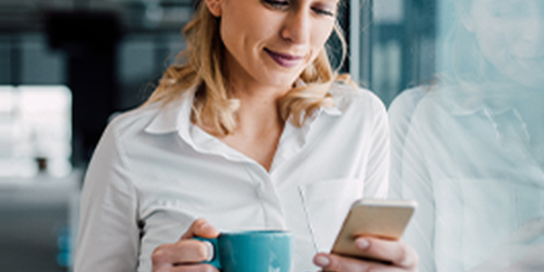 A woman in a white shirt is holding a blue mug and looking at her smartphone. She stands near a window in a modern, light-filled office environment.