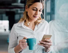 A woman in a white shirt is holding a blue mug and looking at her smartphone. She stands near a window in a modern, light-filled office environment.