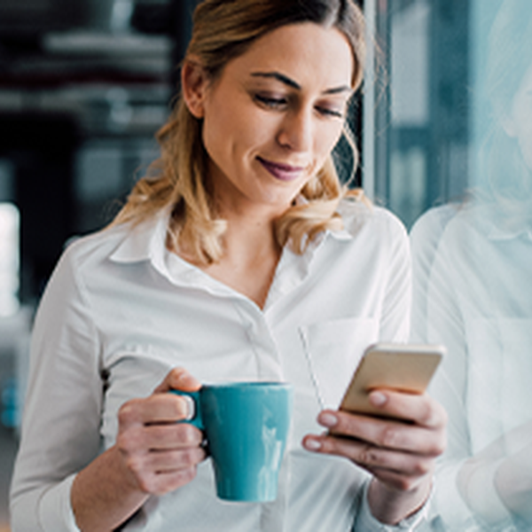 A woman in a white shirt is holding a blue mug and looking at her smartphone. She stands near a window in a modern, light-filled office environment.
