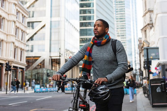 A man in a gray sweater and colorful scarf stands beside a bicycle, looking around in an urban setting with modern skyscrapers and pedestrians in the background.