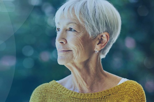 A senior woman with short gray hair gazes thoughtfully into the distance, wearing a bright yellow sweater. The background features an abstract blur of soft blue and green tones, suggesting a serene outdoor setting.