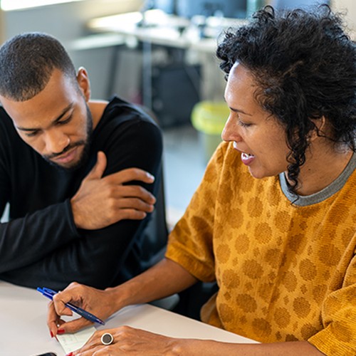 A woman in a patterned orange top writes notes while discussing with a man in a black shirt. They are seated at a table in a bright, modern office environment.