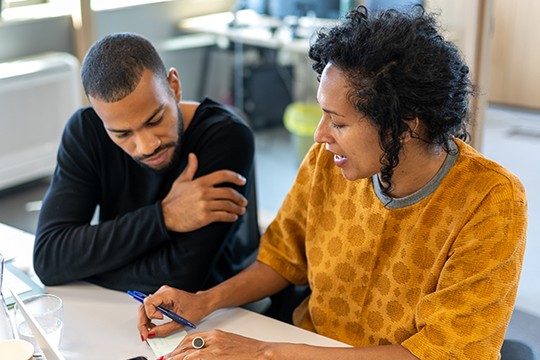 A woman in a patterned orange top writes notes while discussing with a man in a black shirt. They are seated at a table in a bright, modern office environment.
