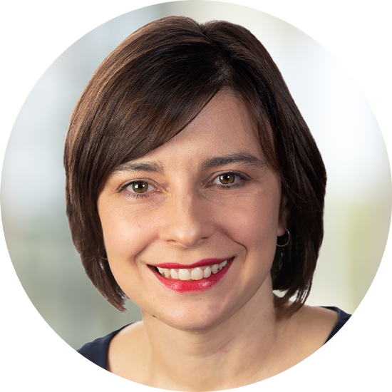 A woman with short brown hair smiles warmly, displaying a bright red lipstick. She appears in a professional setting, with a blurred light backdrop suggesting an indoor environment.