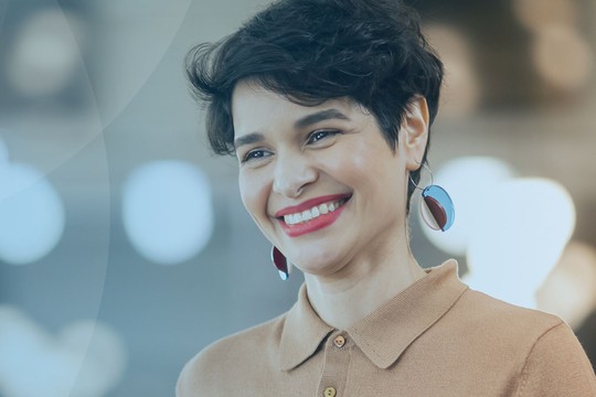 A woman with short, curly hair smiles while wearing a tan collared shirt and colorful earrings. She is in a softly lit environment with blurred lights in the background.