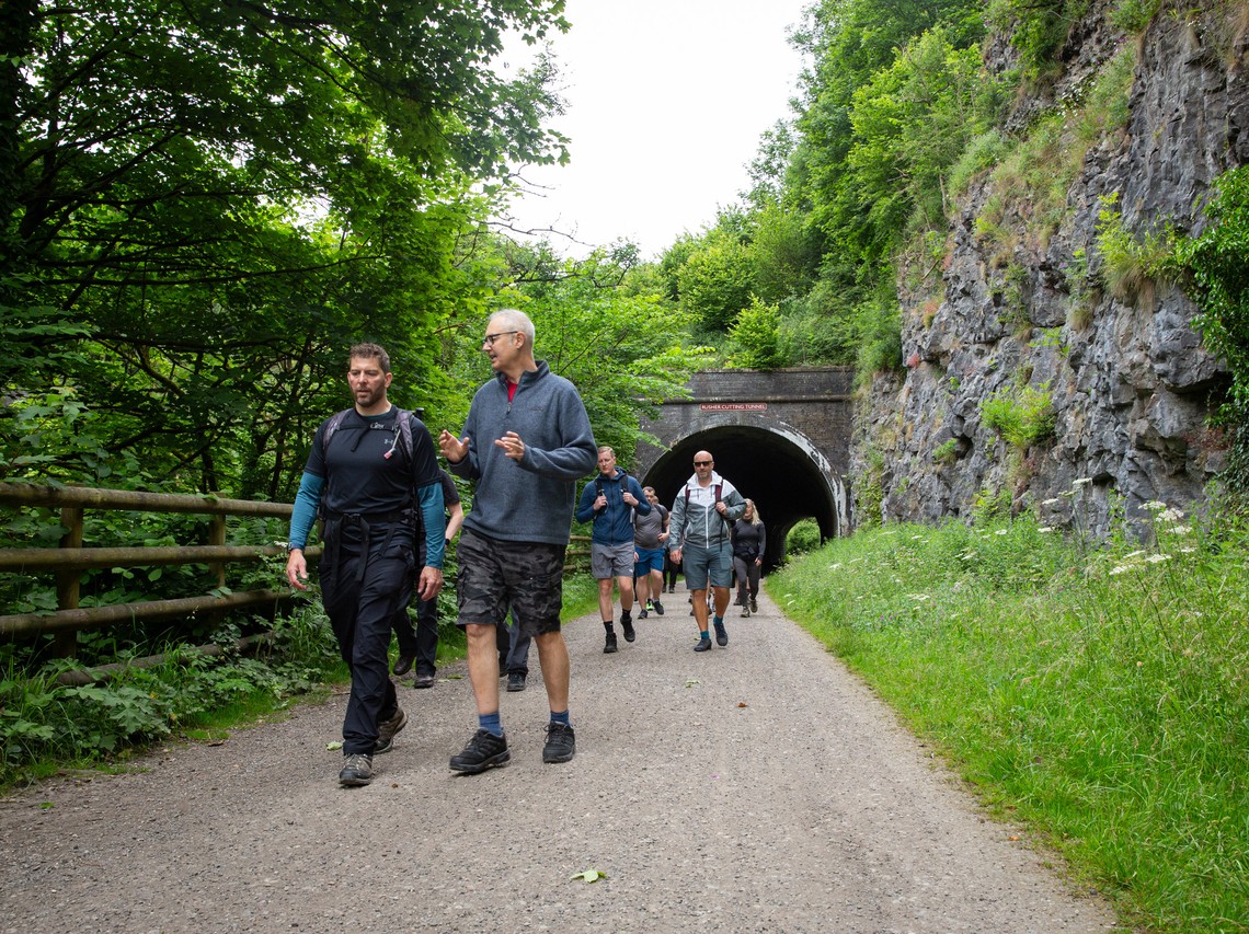 A group of hikers walks along a gravel path surrounded by trees and a rocky hillside, approaching a dark tunnel that leads further into the landscape.