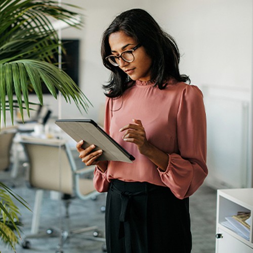 A woman with glasses stands holding a tablet, focused on her device. She wears a pink blouse and black pants, surrounded by a modern office environment with greenery.