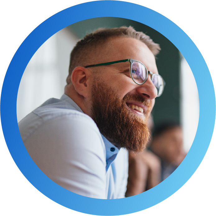 A man with a beard and glasses smiles while seated, engaged in conversation. The setting appears relaxed, with soft lighting and blurred figures in the background.