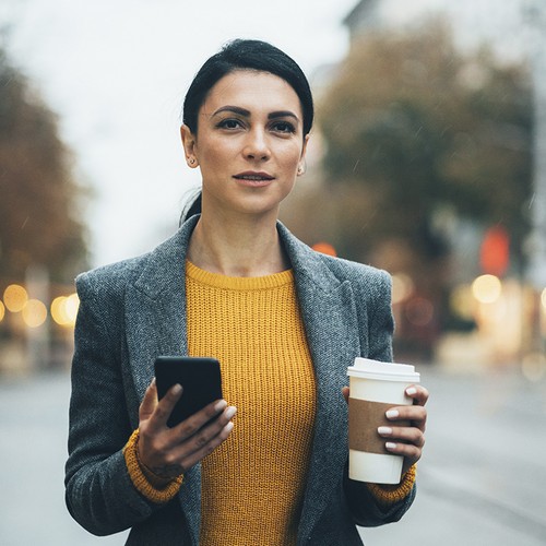 A woman holds a smartphone in one hand and a coffee cup in the other, standing on a city street during a cloudy, rainy day, surrounded by blurred lights and trees.