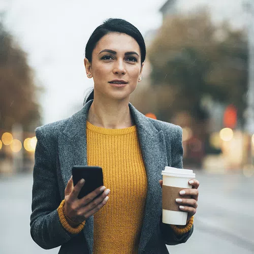 A woman holds a smartphone in one hand and a coffee cup in the other, standing on a city street during a cloudy, rainy day, surrounded by blurred lights and trees.