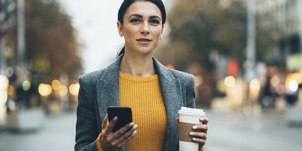 A woman holds a smartphone in one hand and a coffee cup in the other, standing on a city street during a cloudy, rainy day, surrounded by blurred lights and trees.
