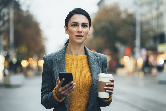 A woman holds a smartphone in one hand and a coffee cup in the other, standing on a city street during a cloudy, rainy day, surrounded by blurred lights and trees.