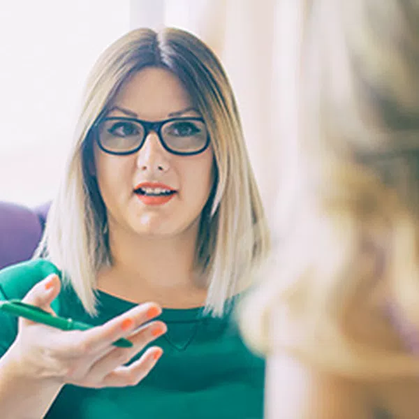 A woman with glasses and shoulder-length blonde hair is engaged in conversation, gesticulating with a pen while sitting in a cozy, softly lit setting. Another person is partially visible.