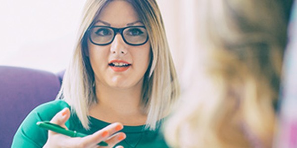 A woman with glasses and shoulder-length blonde hair is engaged in conversation, gesticulating with a pen while sitting in a cozy, softly lit setting. Another person is partially visible.