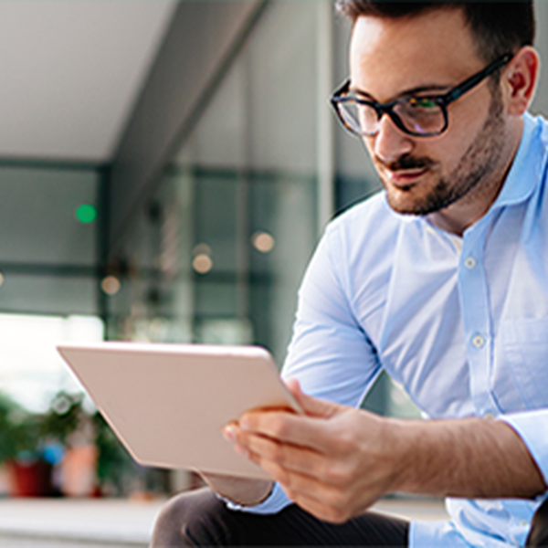 A man in a light blue shirt and glasses sits on steps, intently looking at a tablet. The modern building's glass facade and greenery are visible in the background.