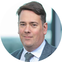 A man with short brown hair wears a grey suit and tie, smiling confidently. He is positioned against a blurred, light-filled background, suggesting a professional or corporate setting.