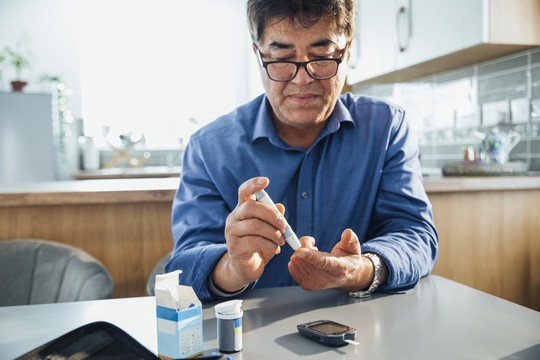 A man in a blue shirt uses a pen-like device to draw blood from his fingertip on a kitchen table, with a glucometer and medication supplies nearby.