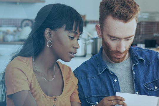 A man and a woman are seated together, focused on a document she's holding. They are in a modern kitchen, surrounded by plants and kitchenware, engaged in a discussion.