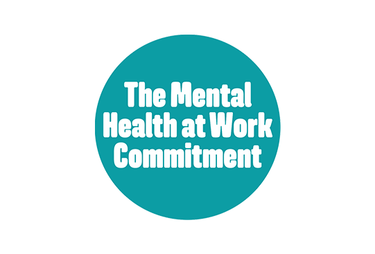 A circular teal badge displays the text “The Mental Health at Work Commitment” in bold, white lettering, indicating a focus on mental health support in workplace environments.