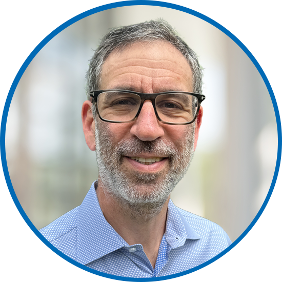 A man with glasses and a beard smiles confidently. He is dressed in a light blue button-up shirt. The background features a soft, blurred environment with natural light.