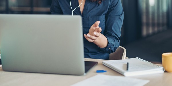 A person wearing a dark shirt gestures while engaging with a laptop at a desk. Nearby items include a notepad, a pen, and a yellow cup, suggesting a workspace setting.
