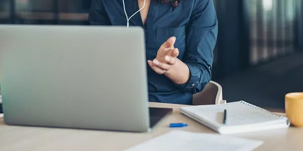 A person wearing a dark shirt gestures while engaging with a laptop at a desk. Nearby items include a notepad, a pen, and a yellow cup, suggesting a workspace setting.