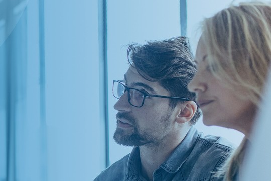 Three individuals are engaged in a discussion, focused on a task. They are in a modern office with large windows, surrounded by natural light reflecting off smooth surfaces.