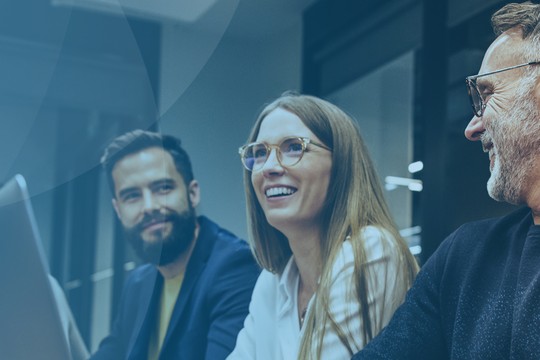 A group of four professionals is engaged in conversation, smiling and sharing ideas in a modern, well-lit office space, with a soft blue backdrop indicating a collaborative atmosphere.