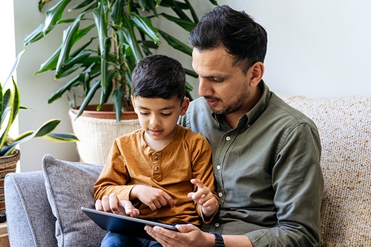 A man and a young boy sit together on a sofa, focusing on a tablet. They are surrounded by indoor plants and a cozy, well-lit living room environment.