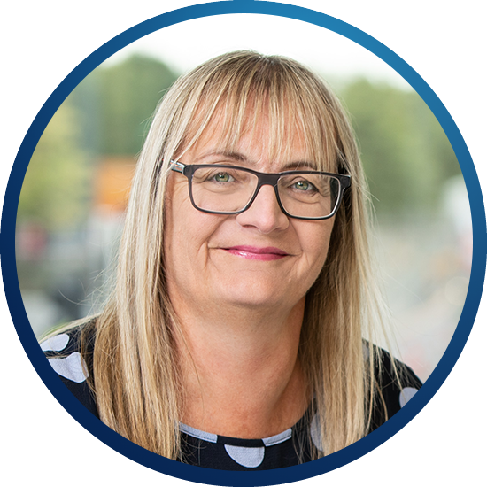 A woman with long straight blonde hair and glasses smiles, framed in a circular crop. She is in a well-lit outdoor setting with blurred greenery and urban elements in the background.