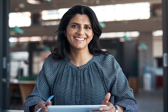 A woman with dark hair smiles while holding a tablet in her hands. She is seated in a modern office setting featuring large windows and a blurred background of workspaces.