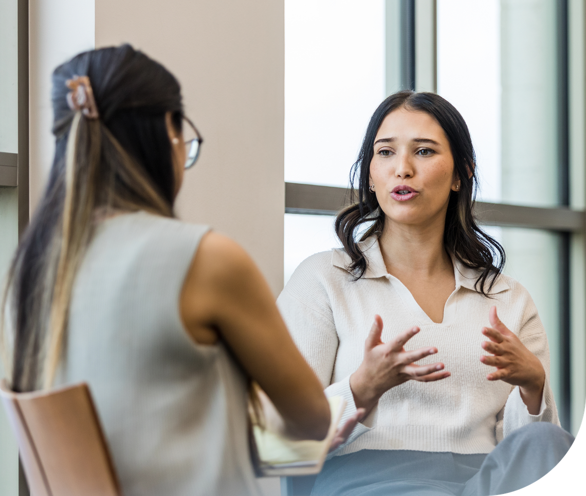 Two women are engaged in conversation. One expresses herself with hand gestures, while the other listens attentively. They sit in a modern, well-lit space near large windows.