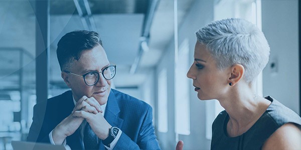 A man in a suit with glasses listens intently to a woman with short hair, gesturing as they engage in a professional conversation in a well-lit office environment.