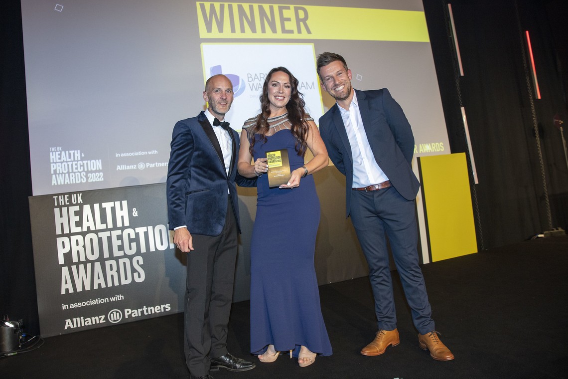 Three individuals stand on a stage, with one woman holding a golden award. They are celebrating a win at the UK Health & Protection Awards 2022, with a backdrop featuring the event's branding.