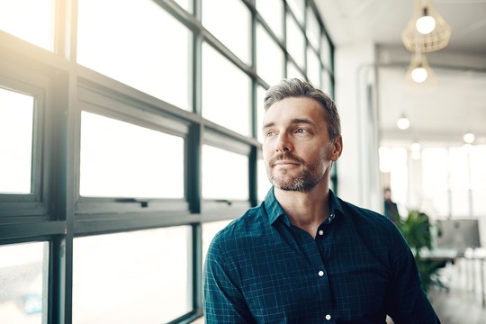 A man with short brown hair and a beard is gazing thoughtfully out of large windows. Soft light enters the modern, sparse workspace filled with greenery and office furniture.