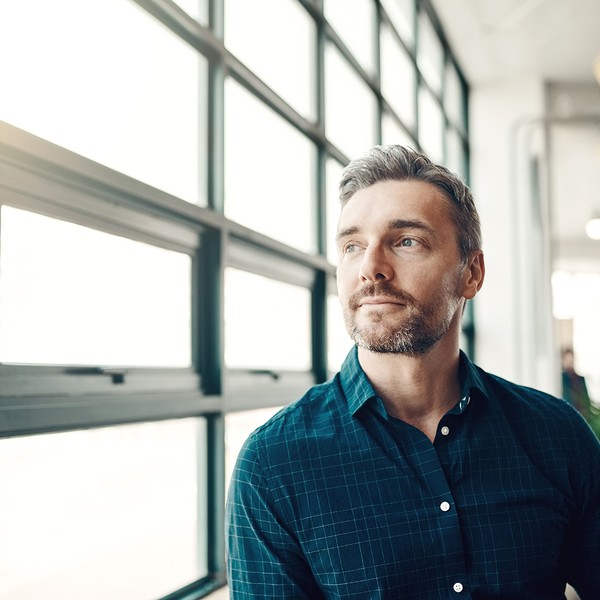 A man with short brown hair and a beard is gazing thoughtfully out of large windows. Soft light enters the modern, sparse workspace filled with greenery and office furniture.